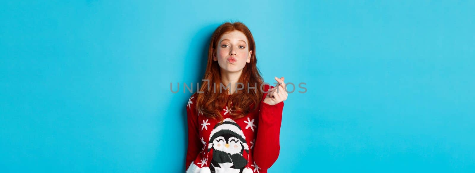 Winter holidays and Christmas Eve concept. Lovely redhead girl in xmas sweater, showing heart sign and pucker lips for kiss, standing over blue background.