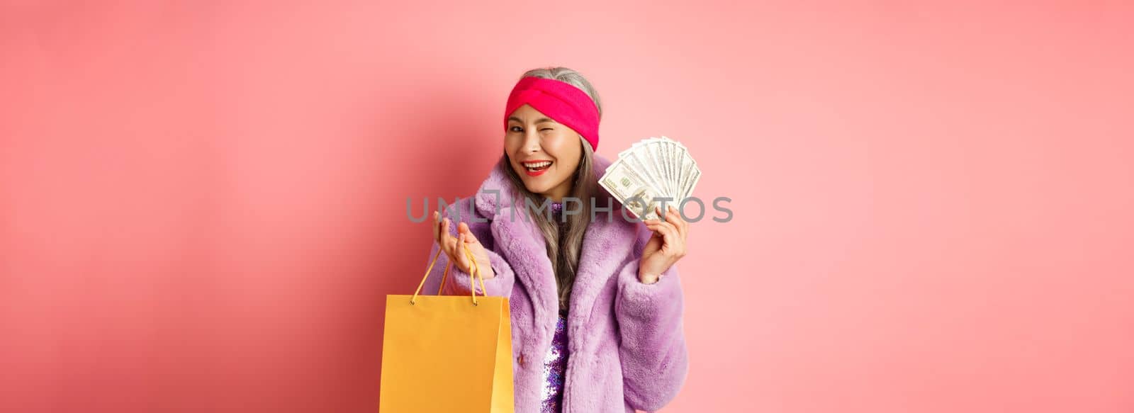 Rich and fashionable asian senior woman wasting money in shops, holding shopping bag and dollars, winking happy at camera, pink background.