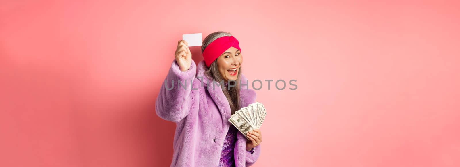 Shopping and fashion concept. Fashionable funky asian woman showing plastic credit card, paying contactless, holding money in other hand, pink background by Benzoix
