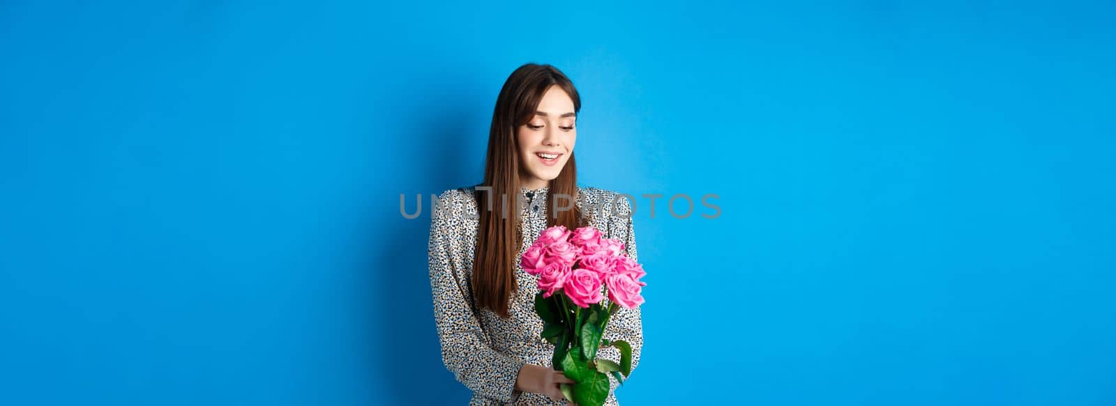 Valentines day concept. Romantic girl in dress looking happy at flowers, smiling at bouquet of pink roses, standing on blue background.