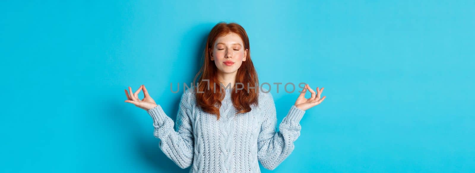 Smiling confident girl with red hair staying patient, holding hands in zen, meditation pose and staring at camera, practice yoga, standing calm against blue background by Benzoix