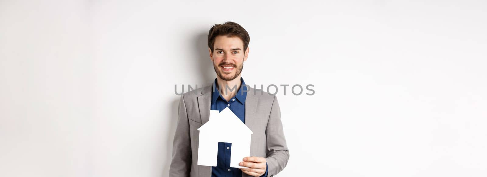 Real estate. Handsome young businessman buying property, holding paper house cutout and smiling, agency advertisement, white background by Benzoix
