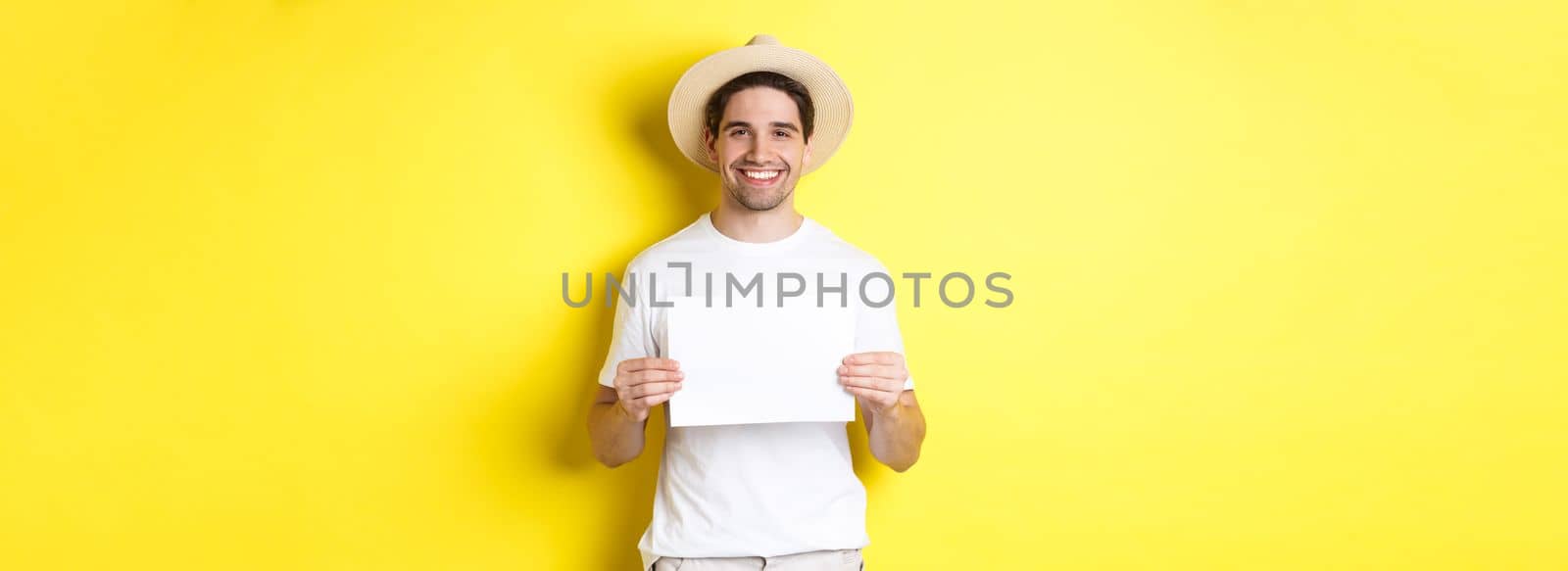 Handsome young male tourist in summer hat smiling, holding blank piece of paper for your sign, standing over yellow background.