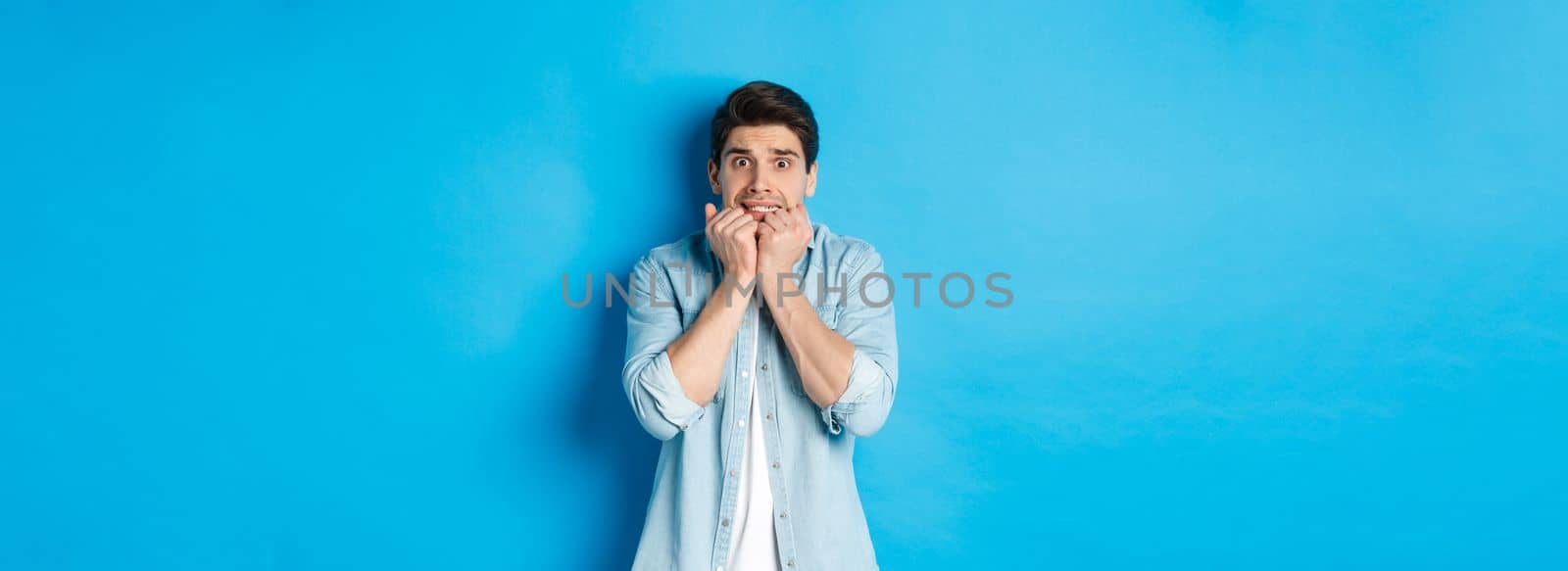 Scared adult man looking at something scary, biting fingernails and trembling from fear, standing over blue background by Benzoix