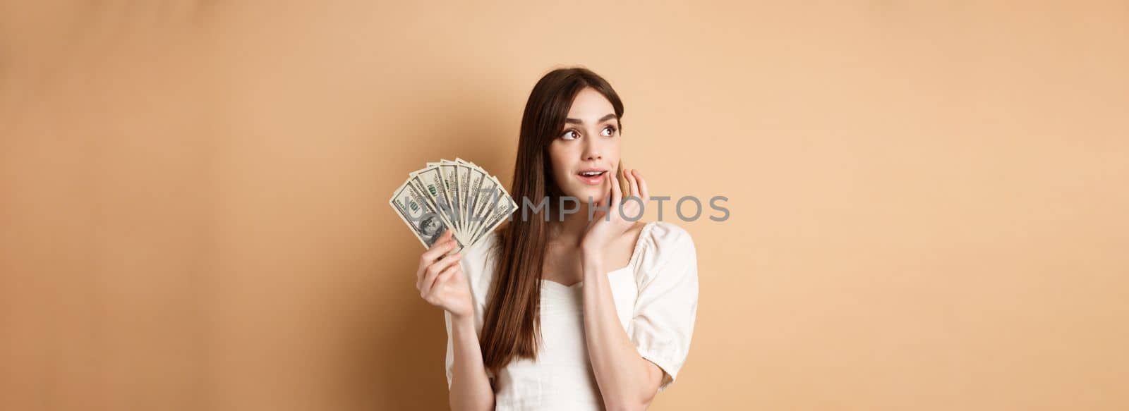 Dreamy girl holding dollar bills and thinking off shopping, looking at upper left corner thoughtful, plan how to waste money, standing on beige background by Benzoix