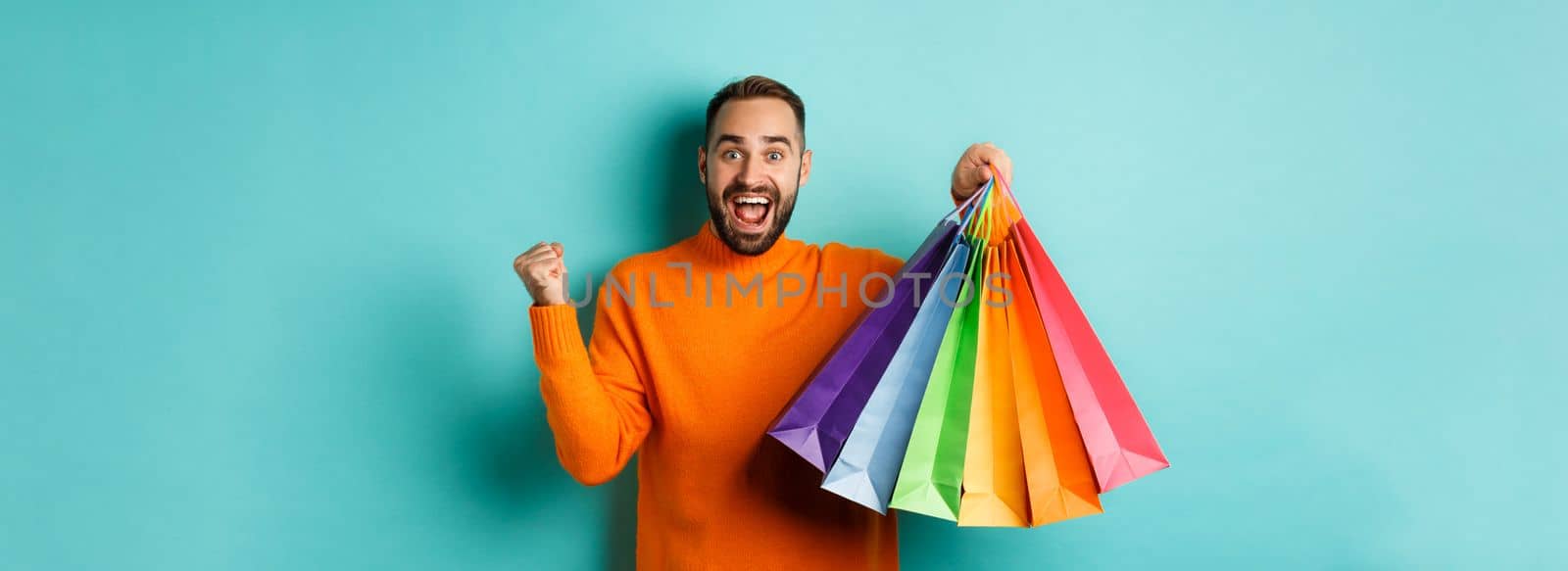 Happy handsome man holding shopping bags and smiling, feeling excitement from discounts, making fist pump, standing over turquoise background by Benzoix