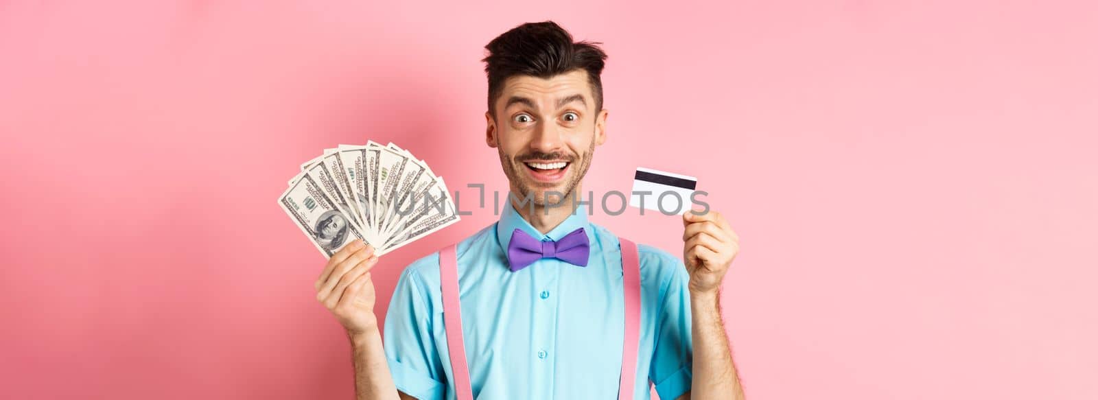 Cheerful caucasian man with moustache and bow-tie showing plastic credit card with money in dollars, smiling at camera, standing over pink background.