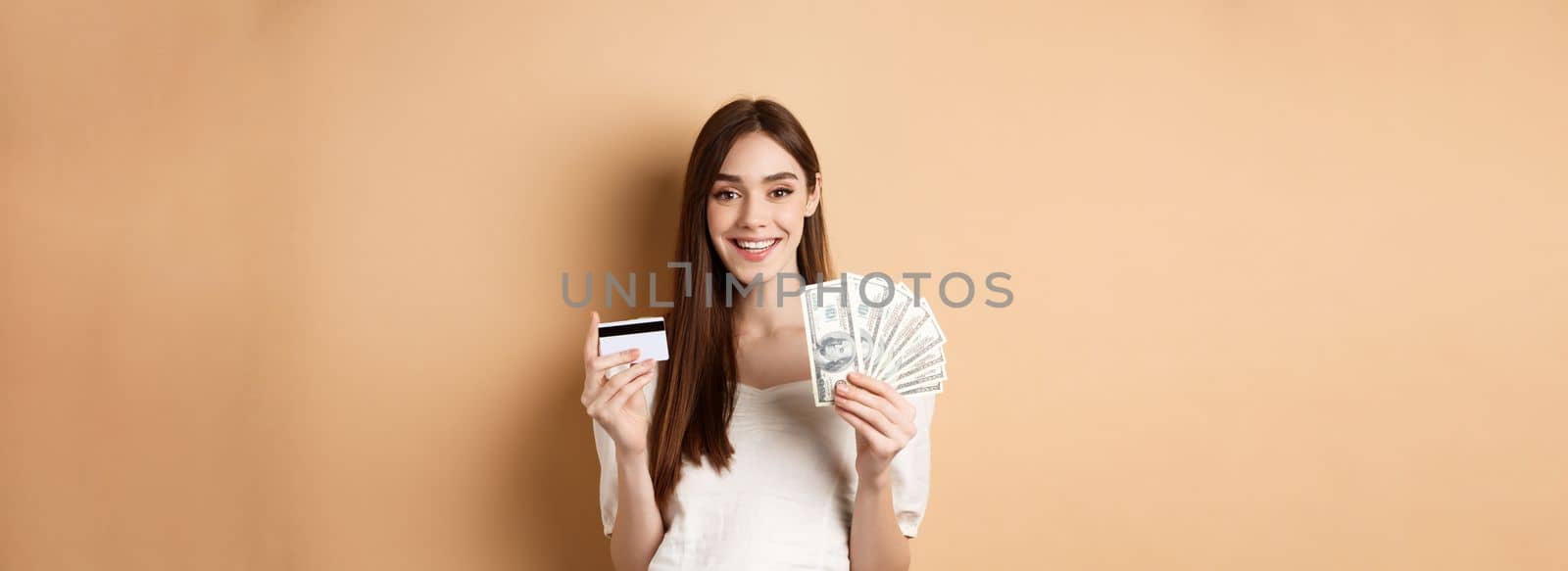 Happy young woman showing dollar bills and plastic credit card, smiling pleased, making money and shopping, standing on beige background.