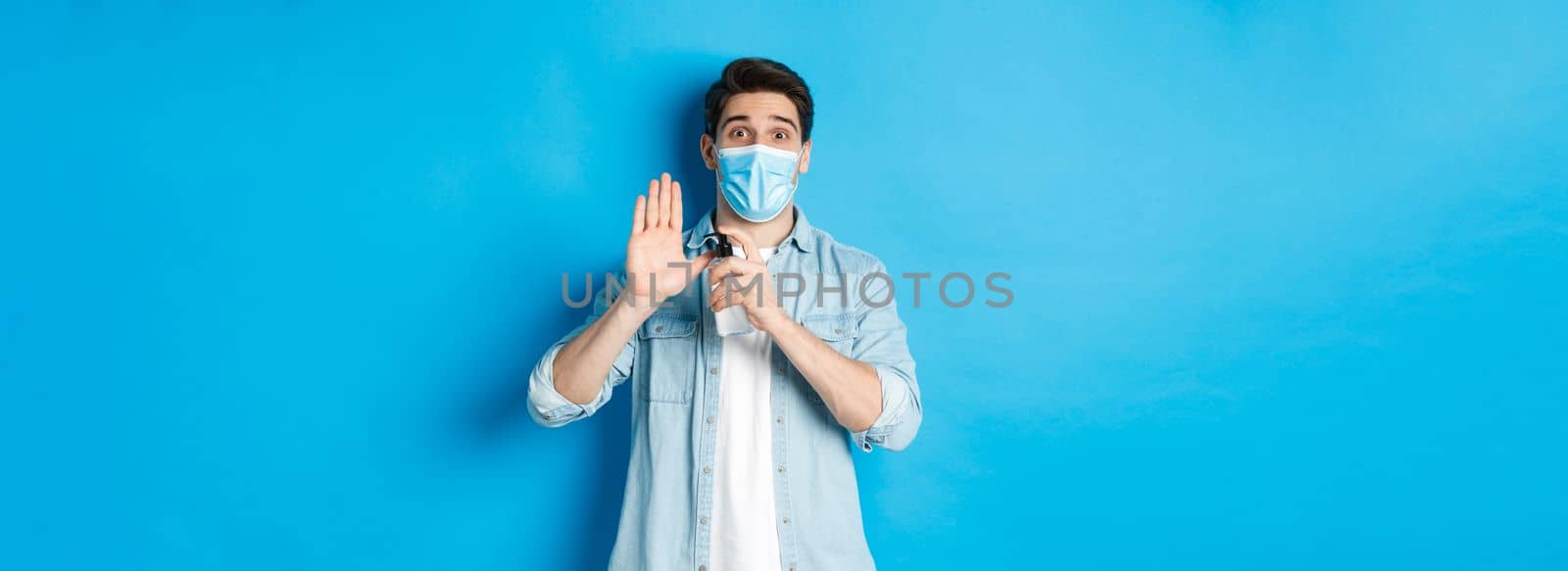 Concept of covid-19, pandemic and social distancing. Cheerful guy in medical mask showing how to disinfect hands with sanitizer, using antiseptic, preventing virus spread, blue background.