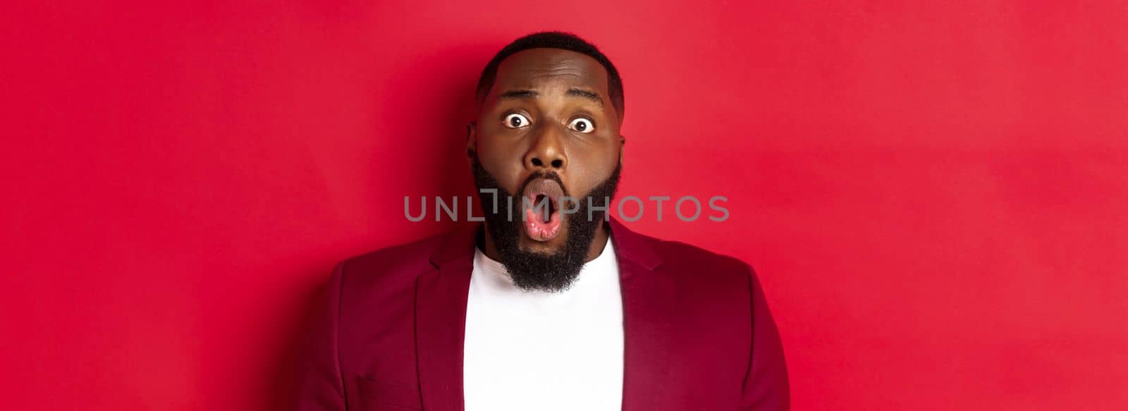 Close-up of shocked Black man gasping and dropping jaw impressed, staring at camera, standing over red background.