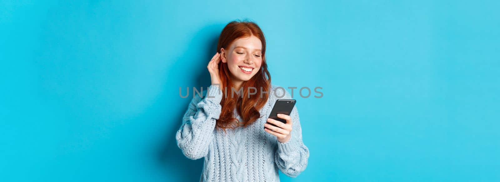 Technology. Happy redhead girl looking pleased at mobile phone, reading compliment in message, smiling and tuck hair behind ear, standing in sweater over blue background.