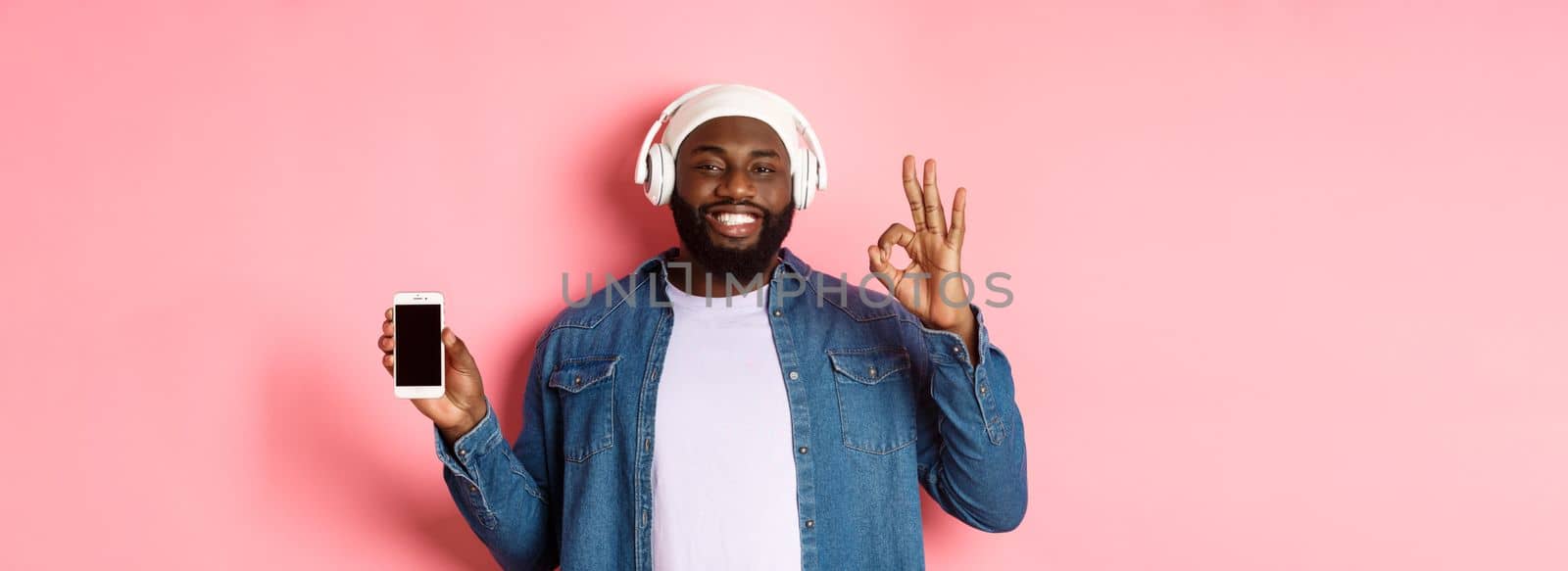Happy hipster guy listening music on headphones and showing mobile screen, smiling satisfied, showing okay sign in approval, like playlist, standing over pink background by Benzoix