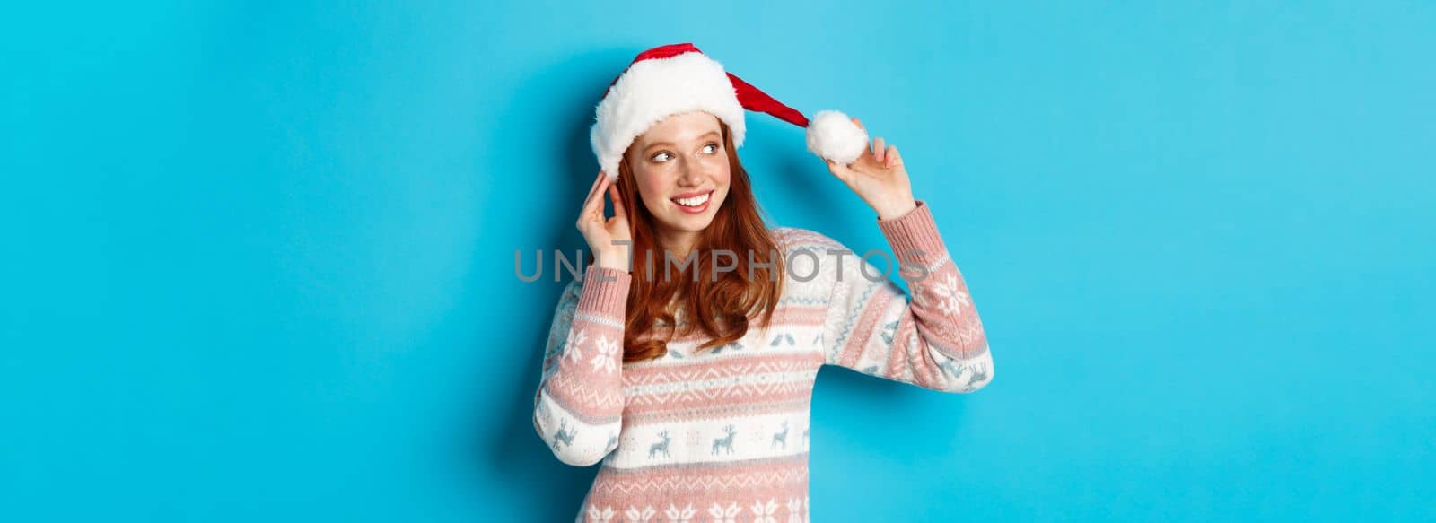 Winter and Christmas Eve concept. Cute teenage girl wearing santa hat and sweater, celebrating xmas, smiling happy, standing over blue background.