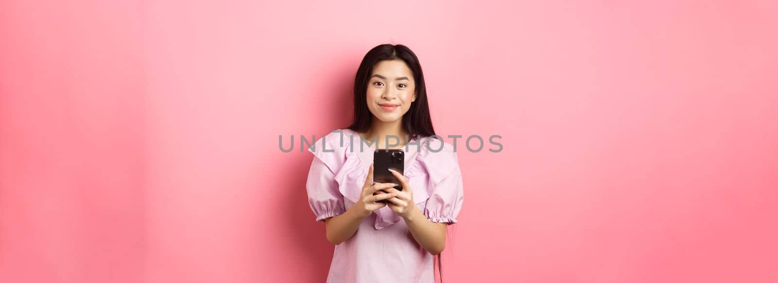 Online shopping. Cute asian girl smiling, holding mobile phone with happy face, standing in dress on pink background.