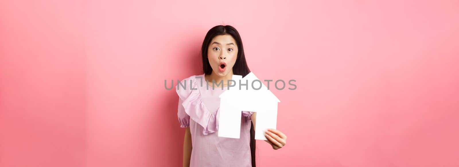 Real estate and insurance concept. Excited asian woman showing paper house cutout and look amazed, special offer, standing on pink background.