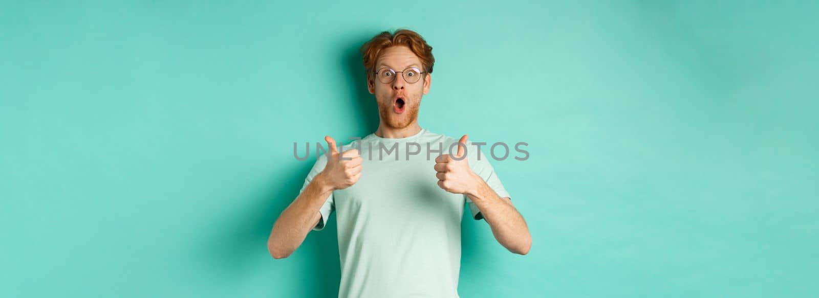 Amazed young man with red hair and beard, wearing glasses with t-shirt, showing thumbs-up and gasping in awe, checking out awesome promo offer, turquoise background.