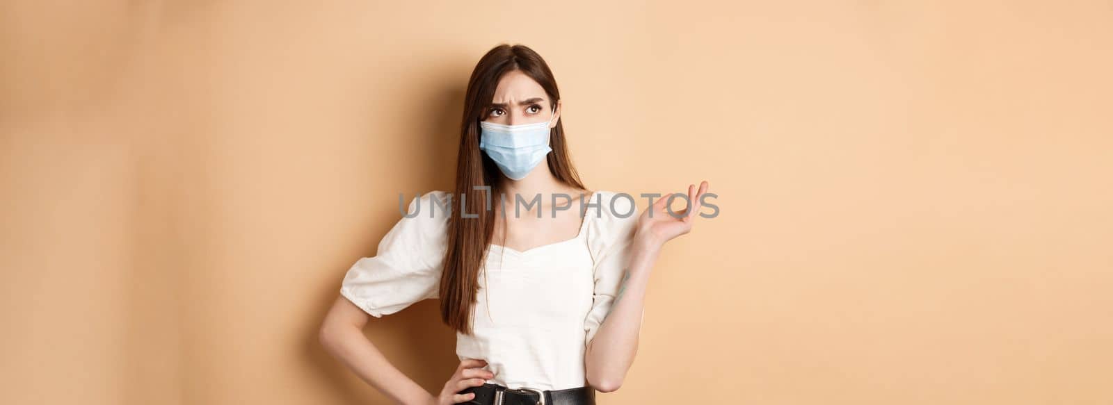Covid-19 and lifestyle concept. Confused girl in face mask looking at upper left corner with troubled face, standing on beige background.