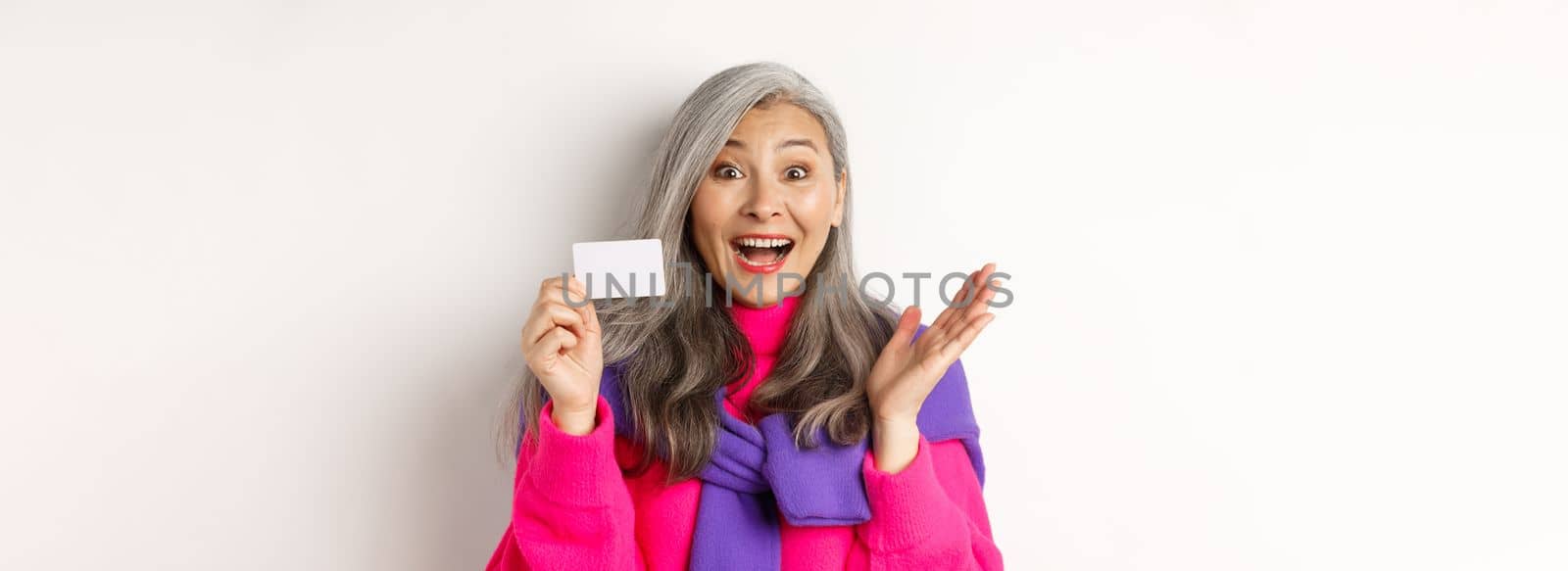 Shopping concept. Happy asian old aldy looking impressed and showing plastic credit card of her bank, standing over white background.
