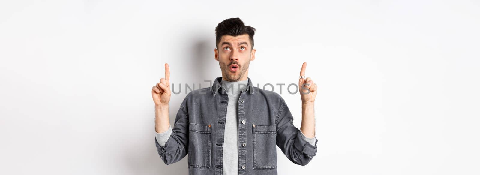 Excited modern man in denim jacket looking and pointing up, gasping amazed, checking out cool offer, standing on white background.