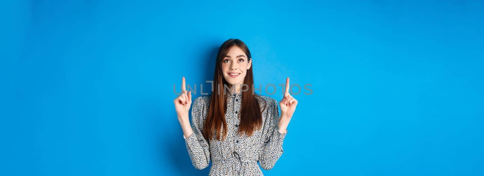 Beautiful and tender young woman pointing fingers up, smiling while advertising promo deal on blue background by Benzoix