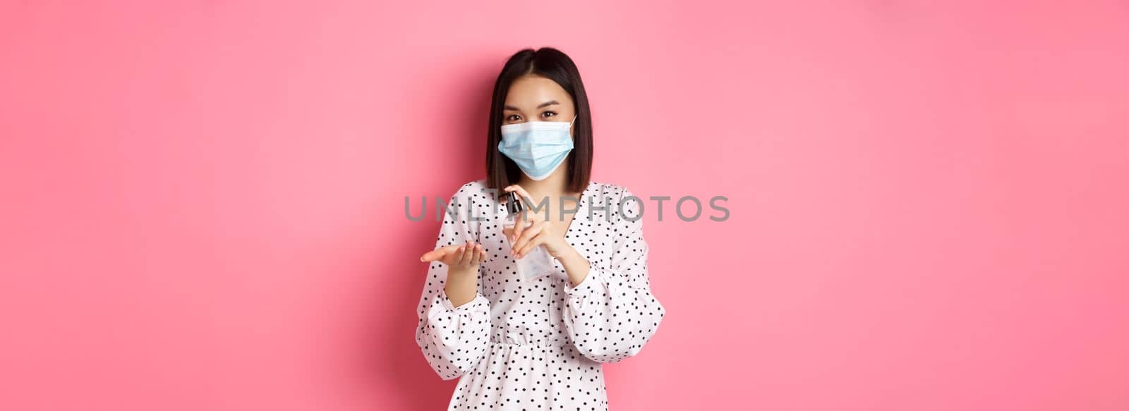 Covid-19, pandemic and lifestyle concept. Cute asian woman clean hands with sanitizer, using antiseptic and wearing medical mask, standing over pink background.