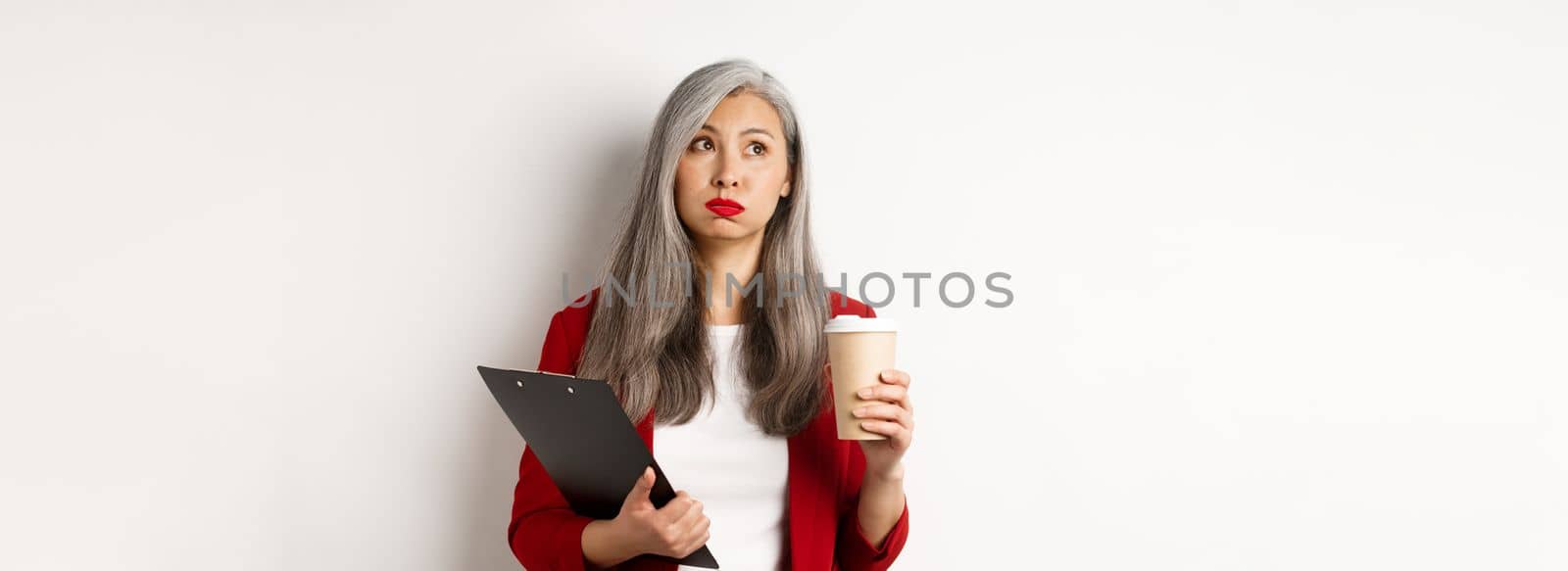 Tired asian female office worker holding clipboard and paper cup, drinking coffee and exhaling with exhausted face, standing over white background.