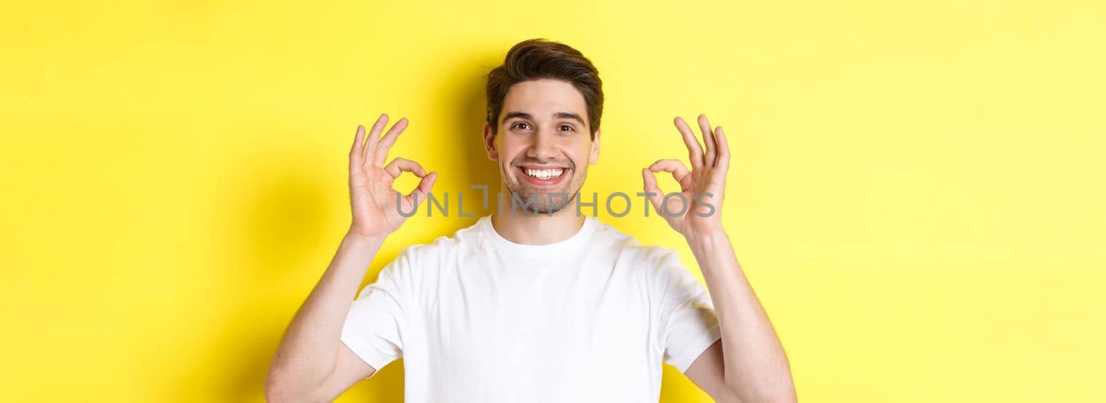 Close-up of handsome young man showing okay sign, approve and agree, smiling satisfied, standing over yellow background by Benzoix