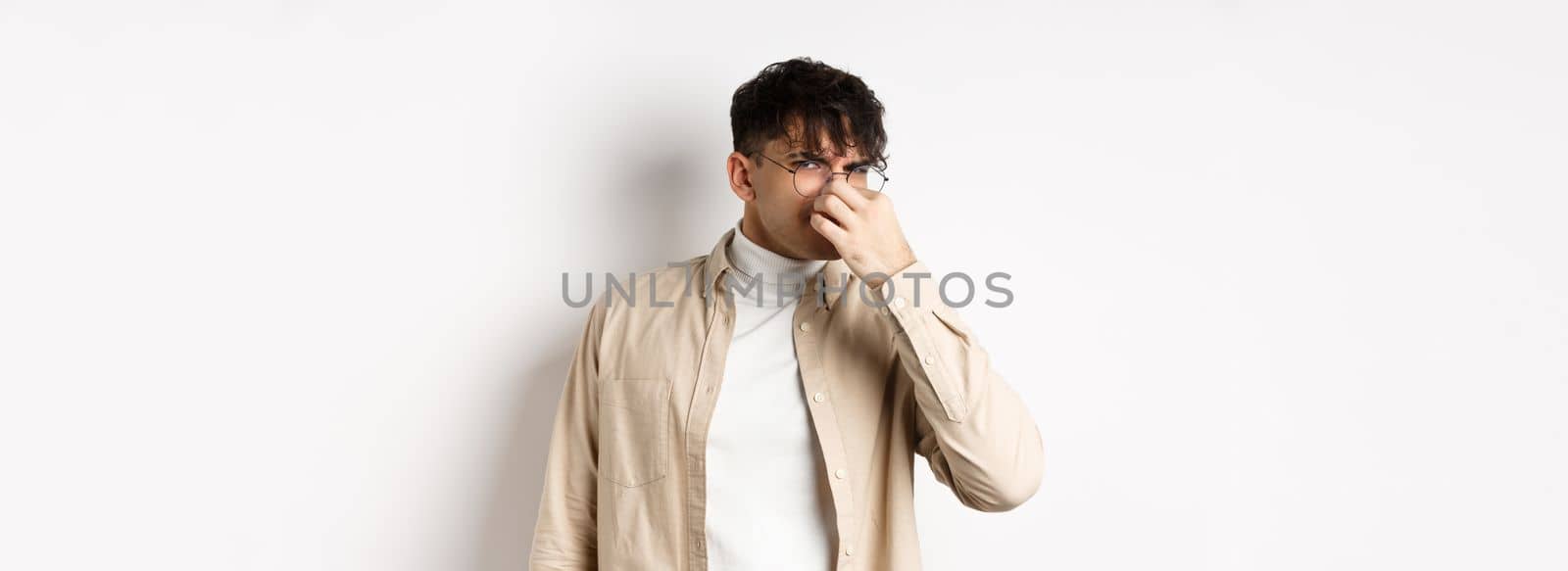 Image of disgusted guy shut his nose from awful smell, looking at something disgusting and stinky, standing on white background.