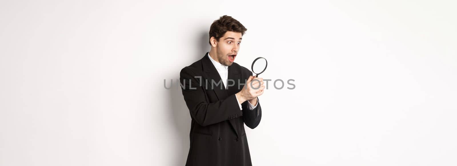 Handsome businessman in black suit, holding magnifying glass and smiling, found something, standing against white background.