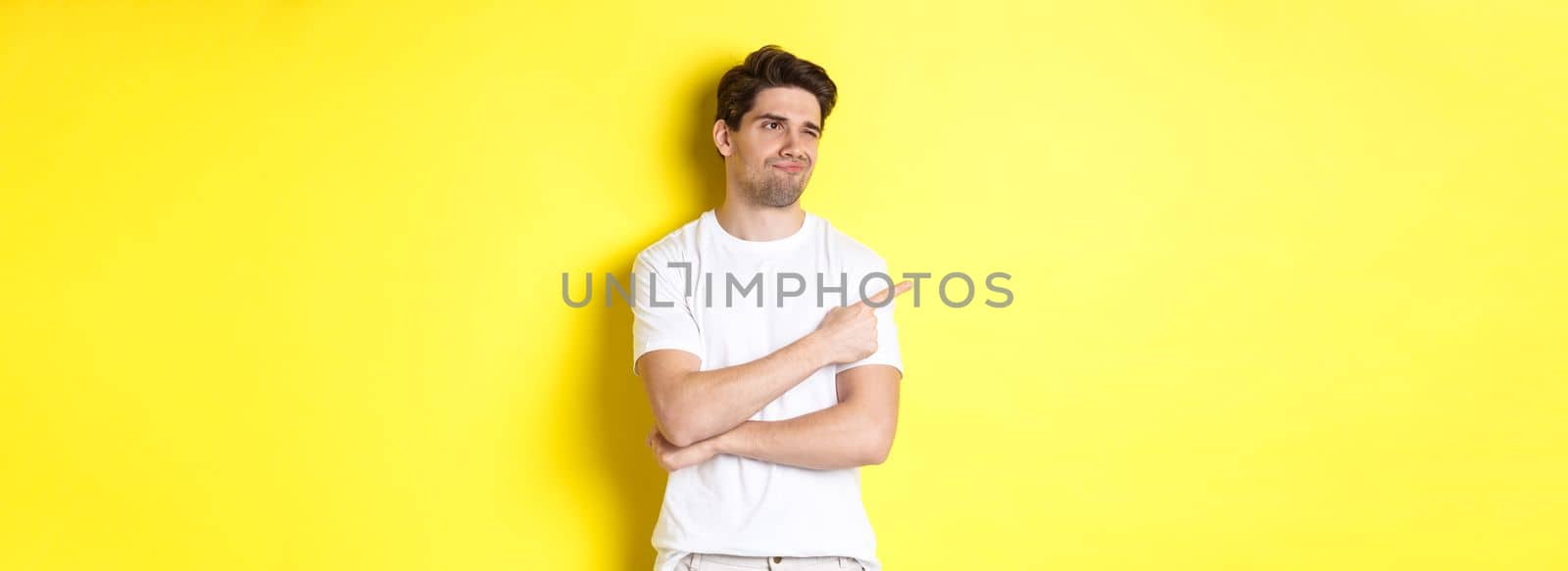 Unimpressed young man complaining, pointing and looking with skeptical face at bad product, standing against yellow background by Benzoix