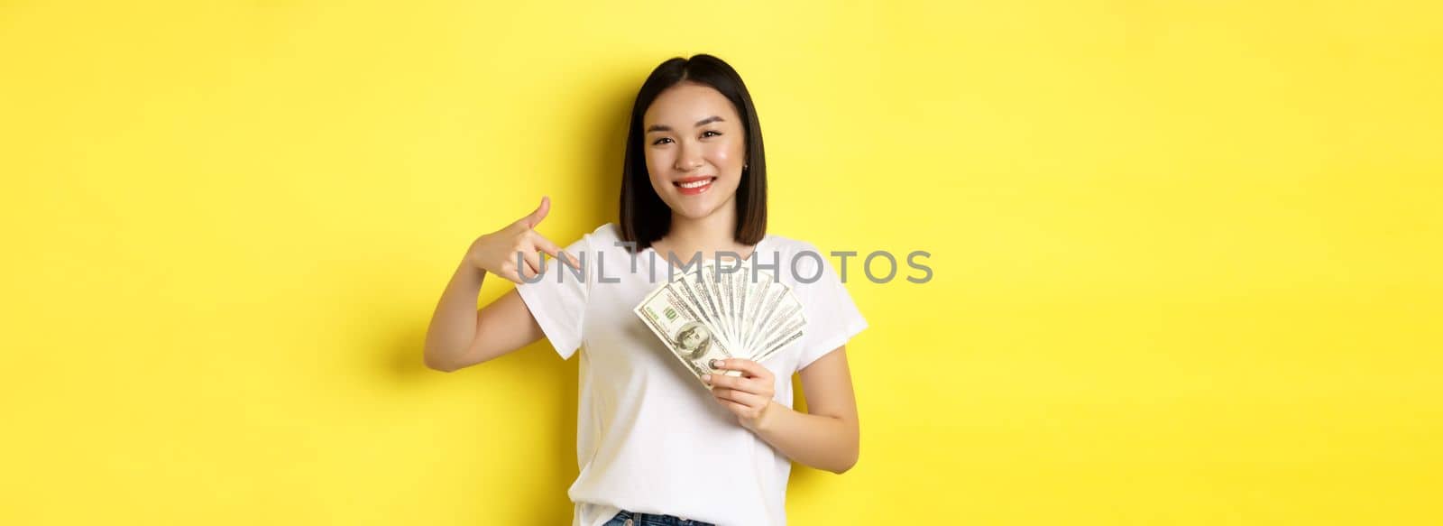 Young asian woman smiling, showing prize money, pointing finger at dollars, standing over yellow background by Benzoix