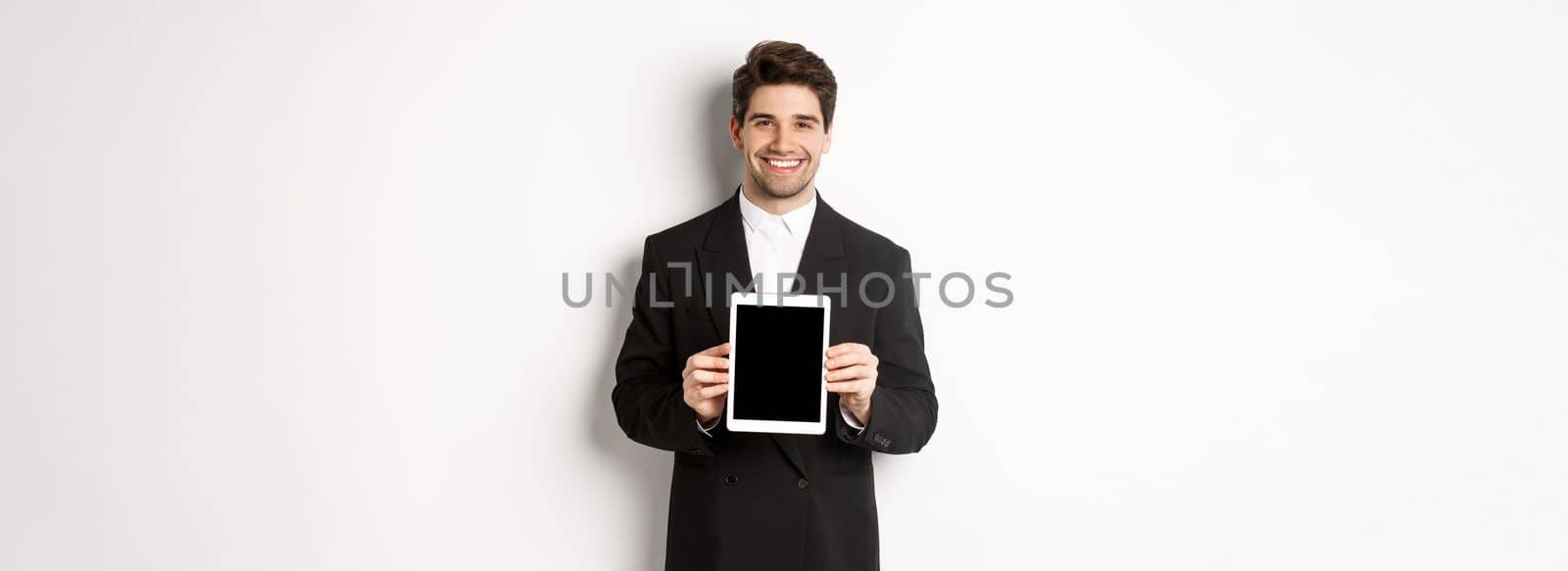 Portrait of handsome businessman in trendy suit, showing digital tablet screen and smiling, standing against white background by Benzoix