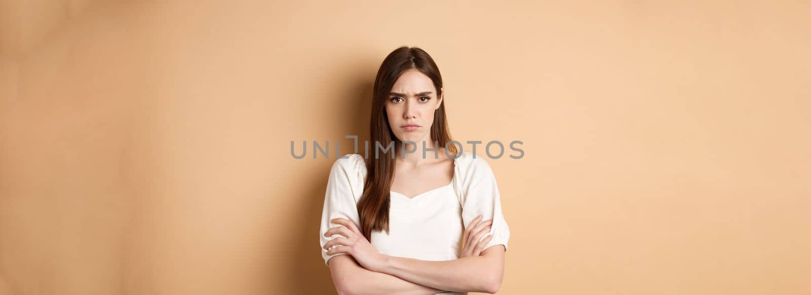 Angry girlfriend cross arms on chest and frowning, looking offended or disappointed, sulking at you, standing mad on beige background.