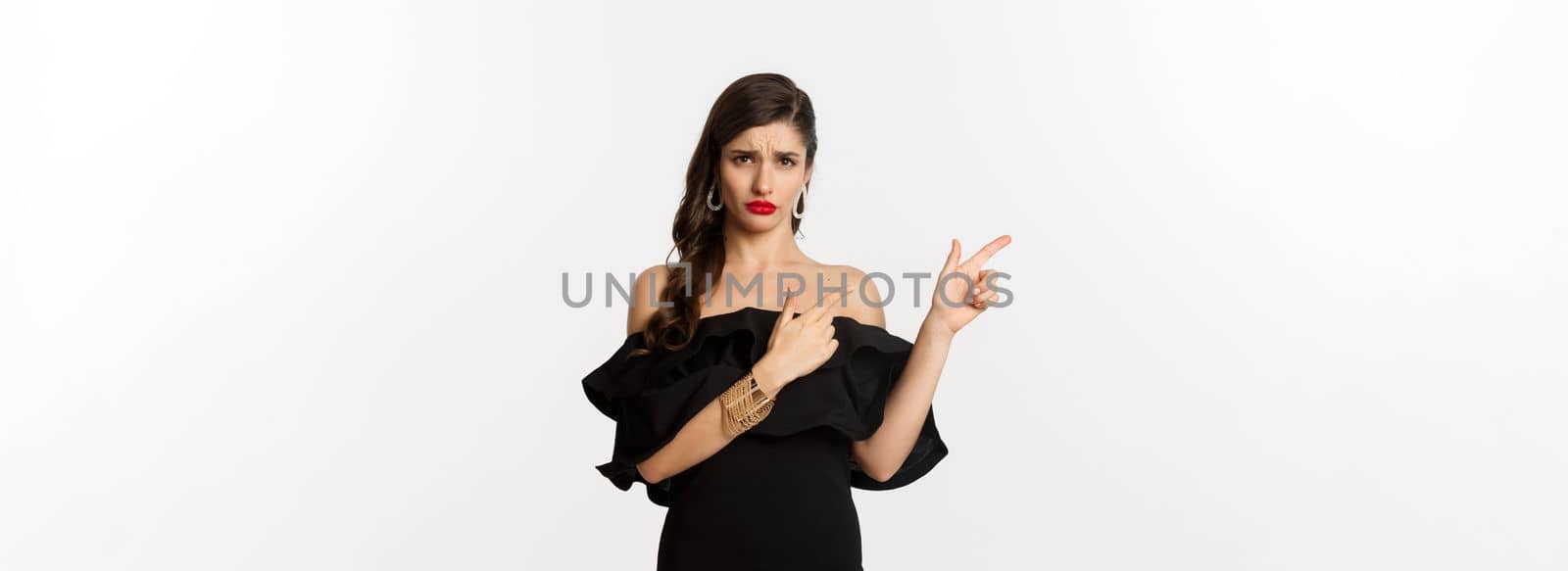 Fashion and beauty. Disappointed glamour woman complaining, wearing black dress, sulking and pointing fingers left at bad offer, white background by Benzoix