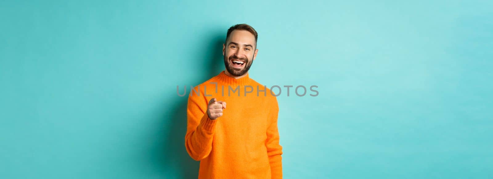 Handsome man laughing and pointing at camera, nodding in approval, agree with you, standing over light blue background.