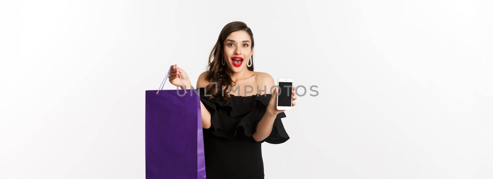 Beauty and shopping concept. Beautiful and stylish woman showing smartphone screen and bag, buying online, standing over white background by Benzoix