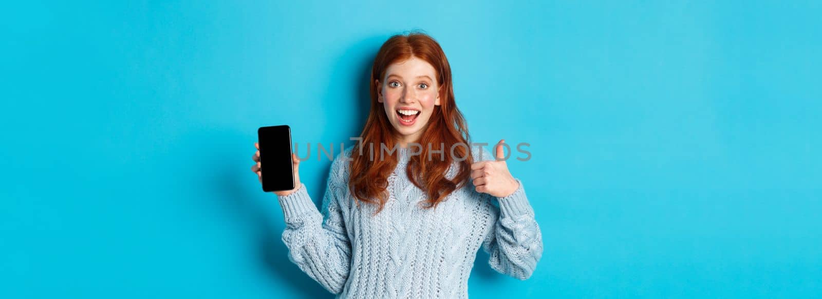 Smiling redhead girl showing smartphone screen, holding phone and demonstrating app, making thumb up in approval, recommending online store, blue background.