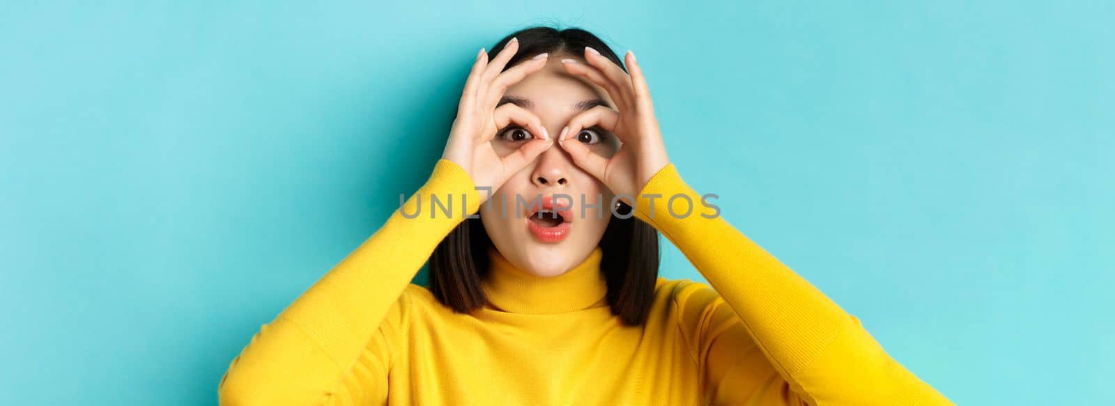 Close up of funny asian woman looking through hand binoculars with surprised face, see something amazing, standing over blue background by Benzoix