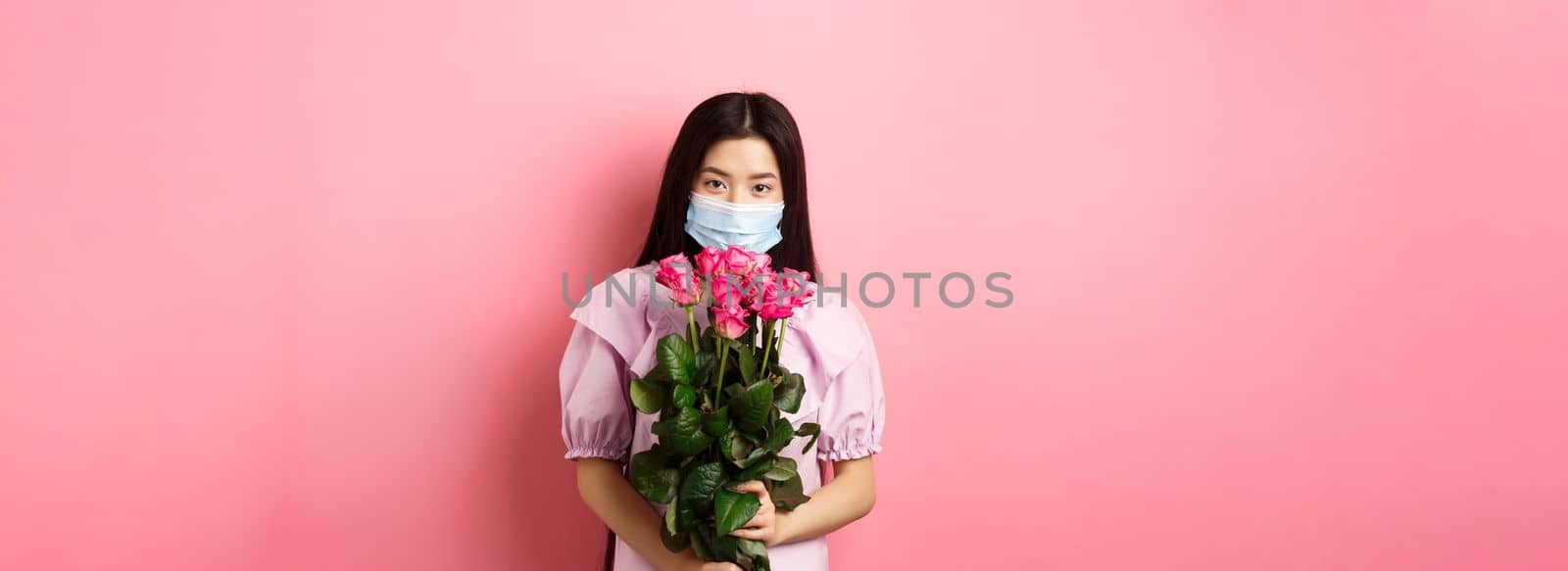 Young asian girl in medical mask holding flowers on Valentines day, receive bouquet of roses from lover, standing on pink background. Social distancing and covid-19 concept.