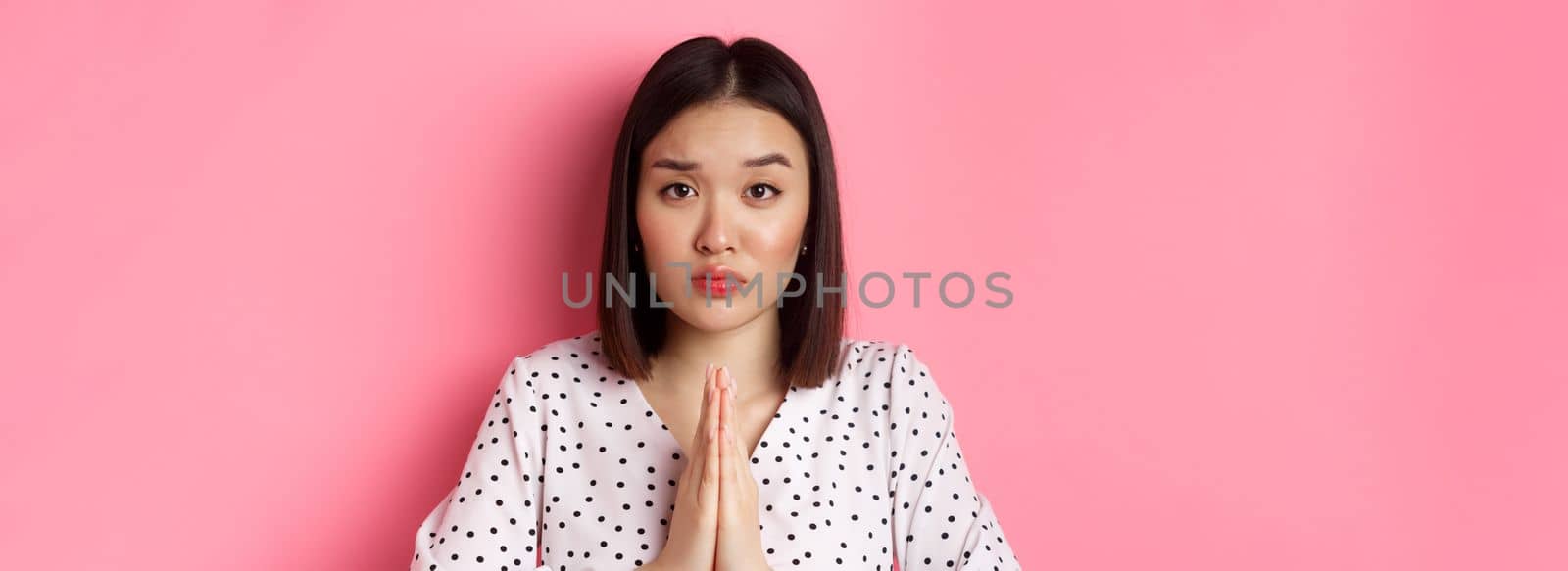 Beauty and lifestyle concept. Sad asian woman asking for help, begging with hands in plead gesture, staring at camera, need favour, standing over pink background by Benzoix