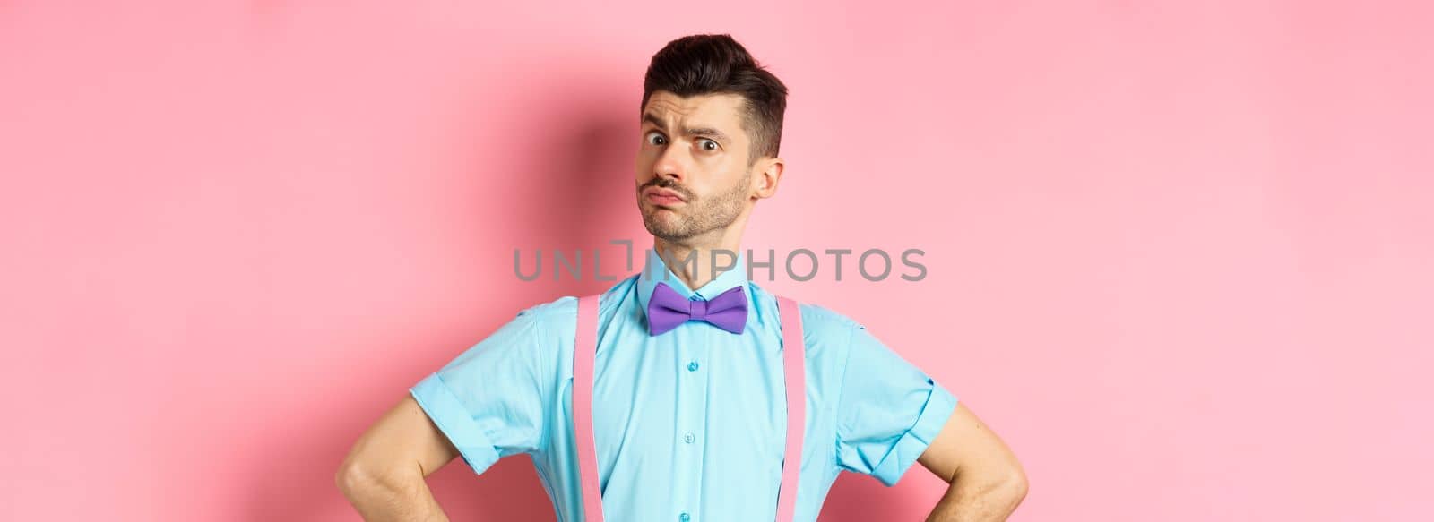 Image of young man with funny moustache and bow-tie standing in confident pose, staring with disbelief and doubts at camera, posing over pink background.