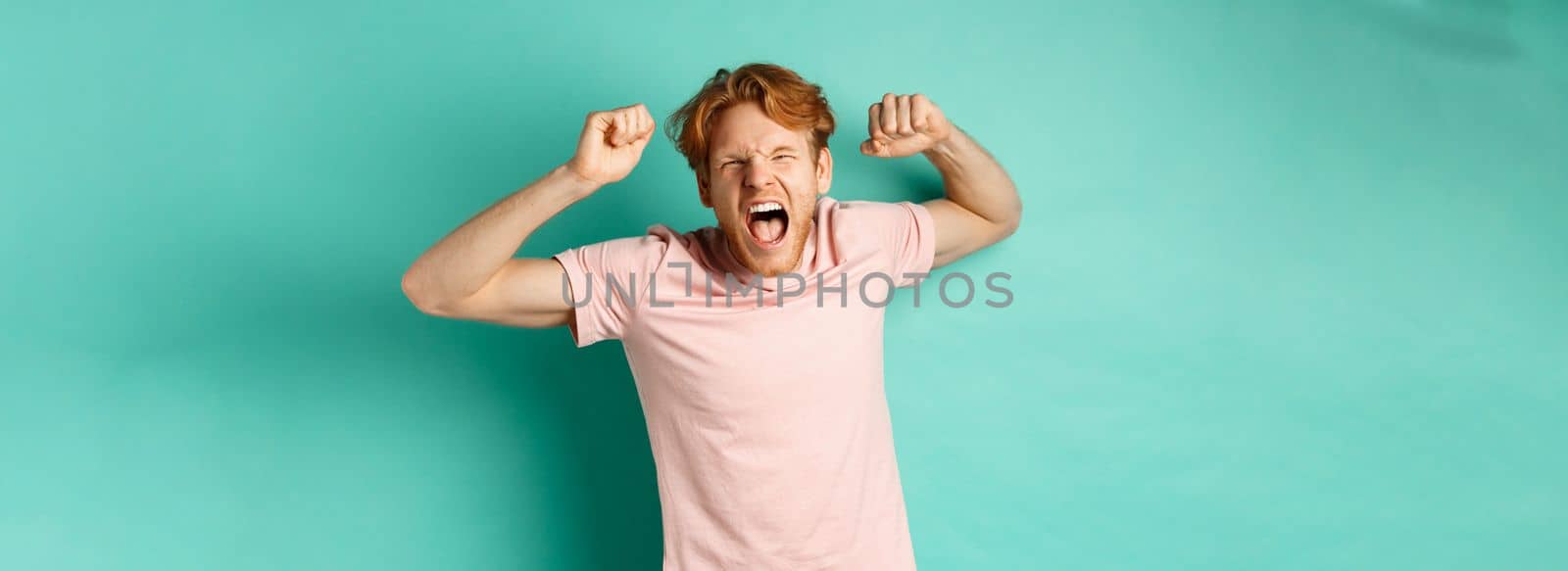 Redhead male fan watching sport game and cheering, shouting and making fist pumps, looking at camera, watching competition, rooting for team, standing over turquoise background.