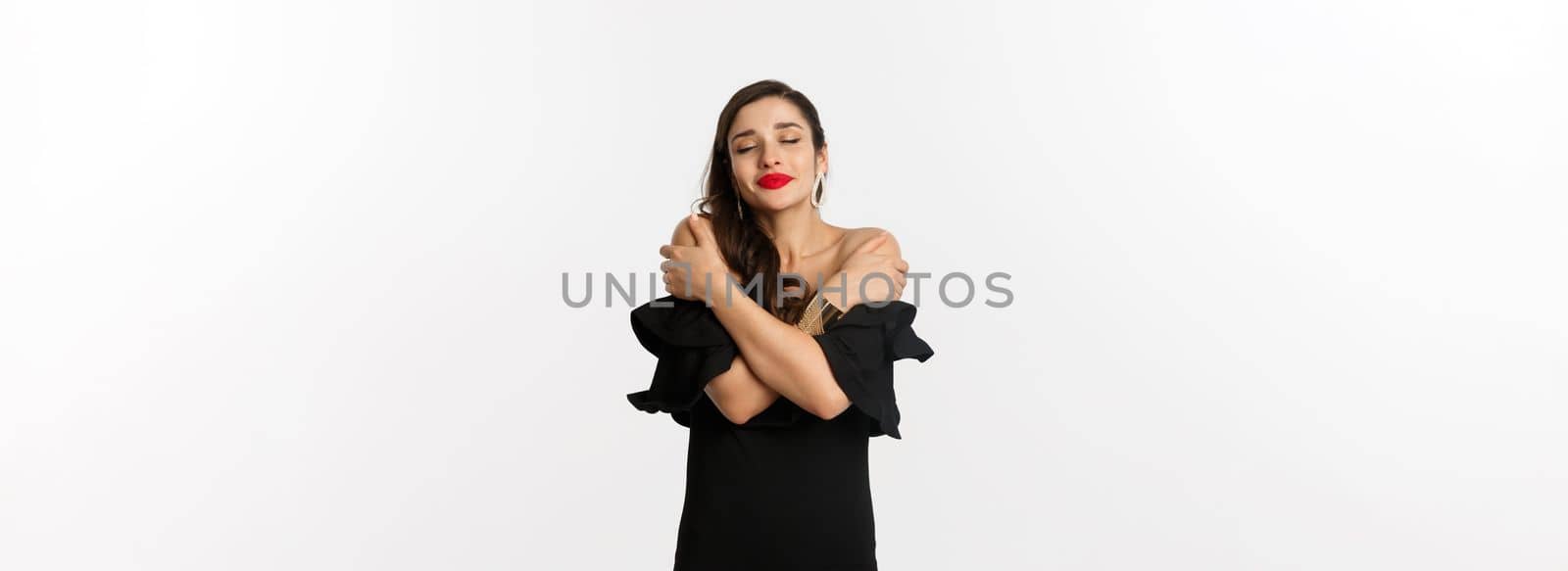 Fashion and beauty. Sensual and beautiful woman in black dress, hugging herself with closed eyes, daydreaming, standing over white background by Benzoix