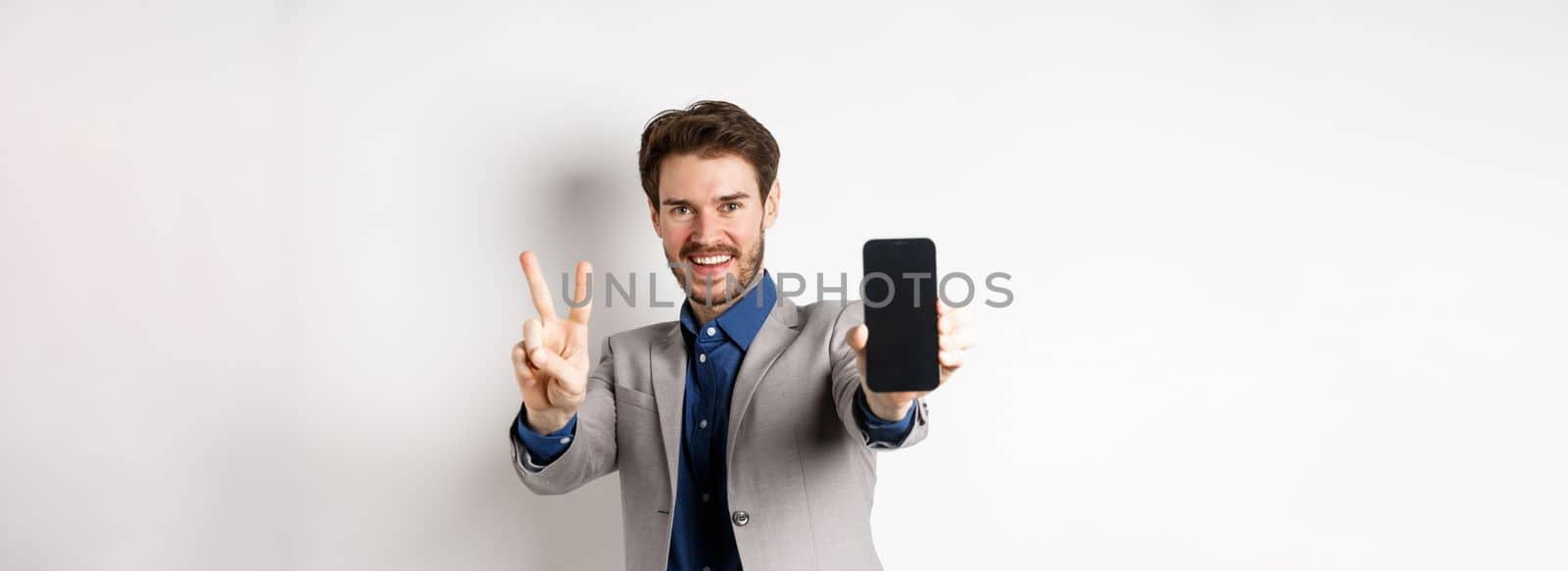 E-commerce and online shopping concept. Cheerful man in business suit showing v-sign and empty mobile phone screen, demonstrate smartphone app, white background by Benzoix