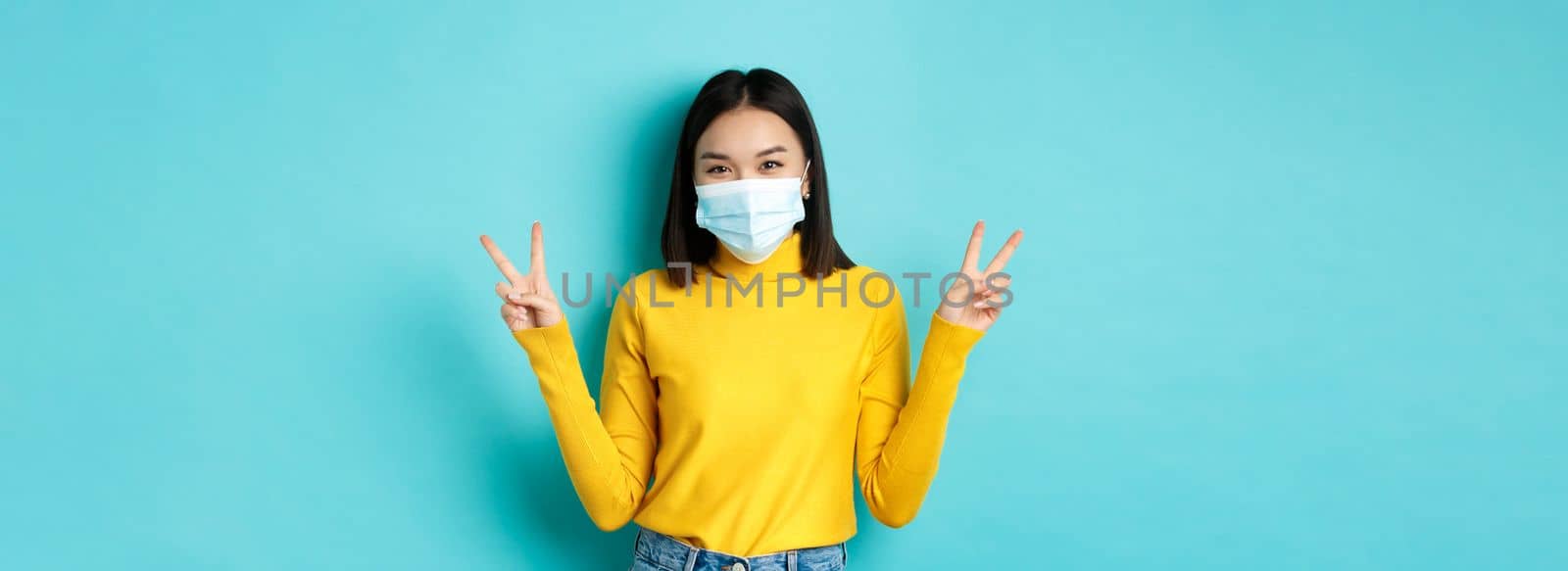 Covid-19, social distancing and pandemic concept. Kawaii asian woman showing peace signs and smiling happy, wearing medical mask from coronavirus disease, standing over blue background by Benzoix