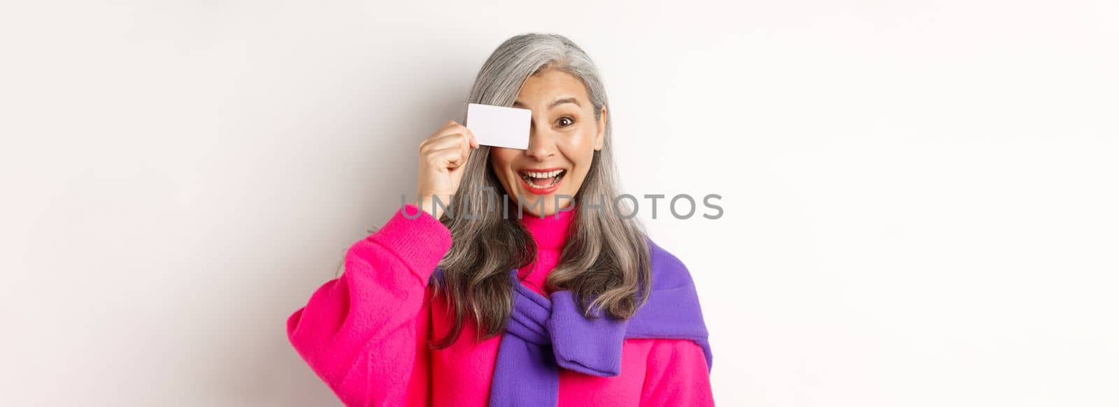 Shopping concept. Stylish asian senior woman smiling and showing plastic credit card, paying contactless, standing over white background.
