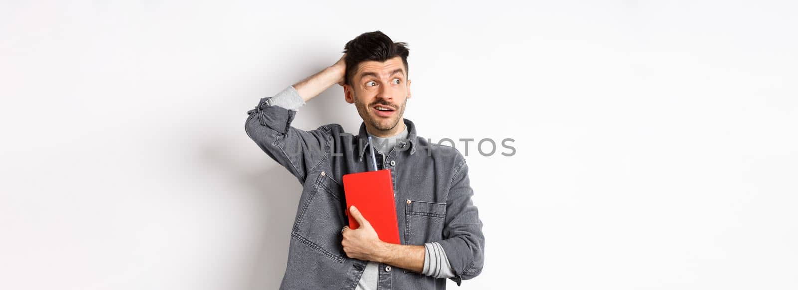 Confused guy scratch head and look aside at logo puzzled, holding red diary or planner, standing against white background.