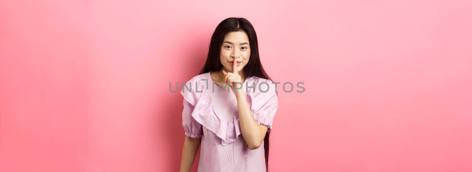 Cute asian girl hiding secret, hushing with finger pressed to lips and smiling, asking to keep quiet, standing in dress on pink background.
