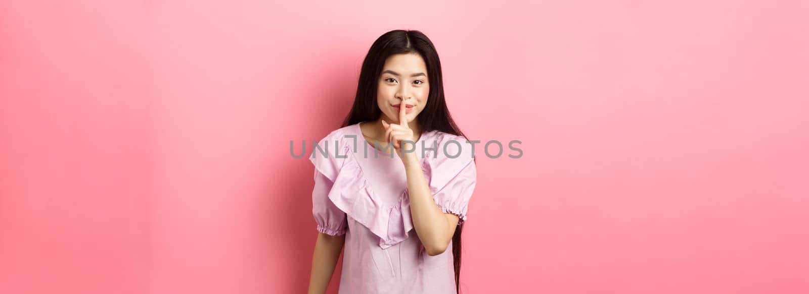 Cute asian girl hiding secret, hushing with finger pressed to lips and smiling, asking to keep quiet, standing in dress on pink background by Benzoix