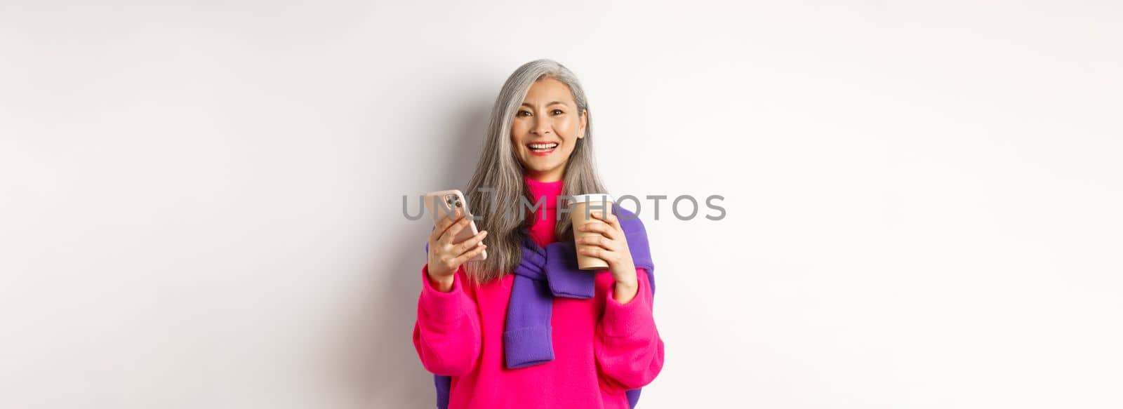 E-commerce concept. Beautiful asian senior woman drinking coffee from takeaway cup and using mobile phone, smiling at camera, white background.
